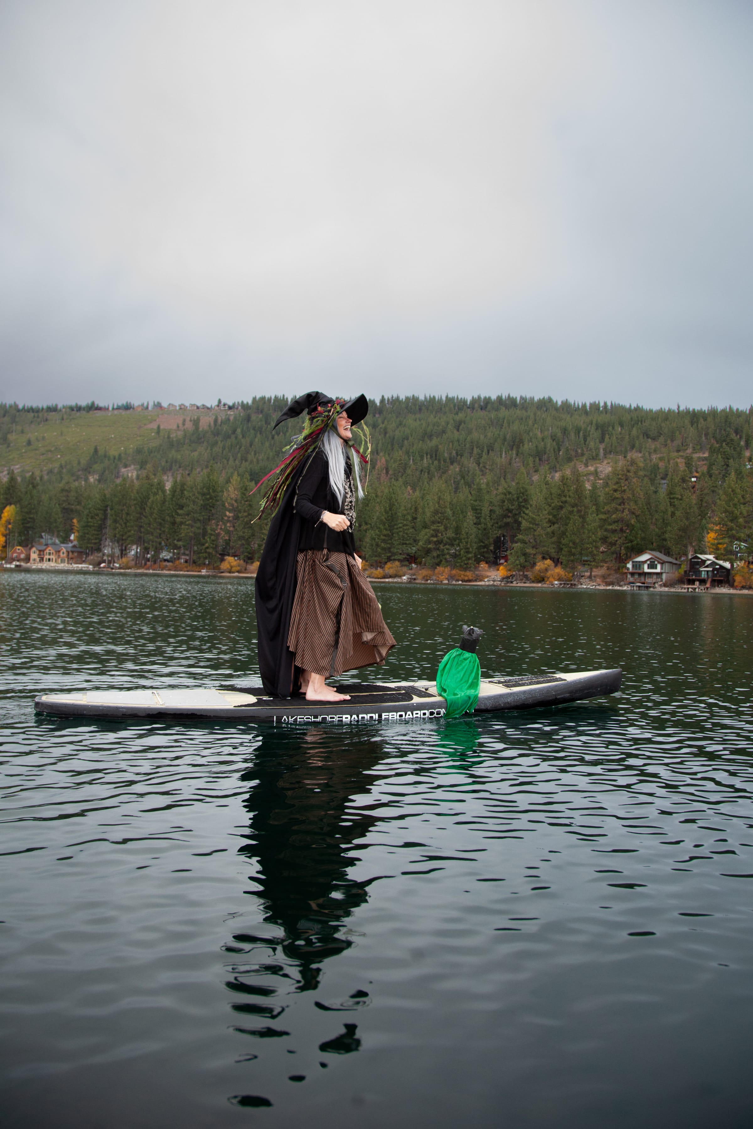 A smiling witch in a flowing hat stands on a paddleboard with a small green familiar.